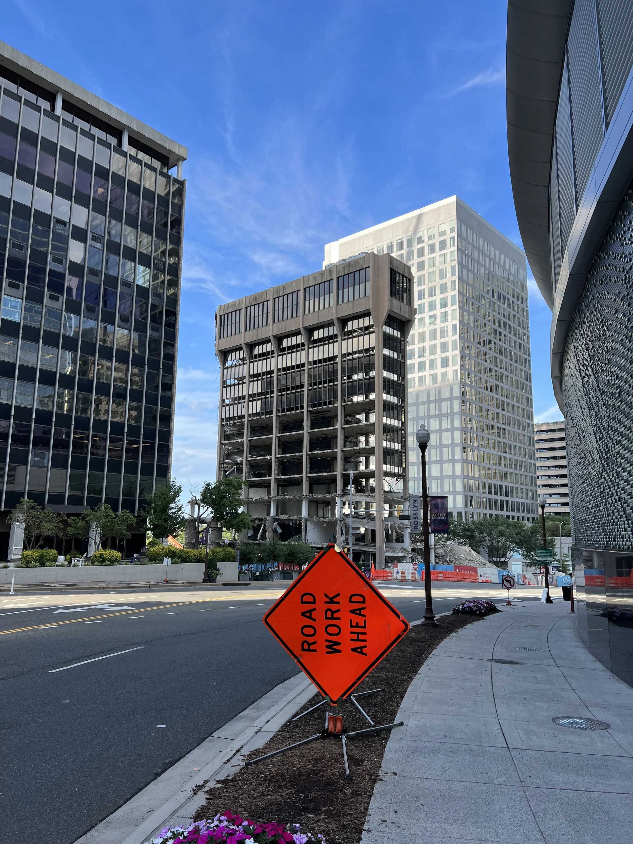 City street with modern buildings, a sidewalk, street lamps, flower beds, and a bright sky. A bright orange roadwork ahead sign is placed on the sidewalk, indicating construction or road work in progress.