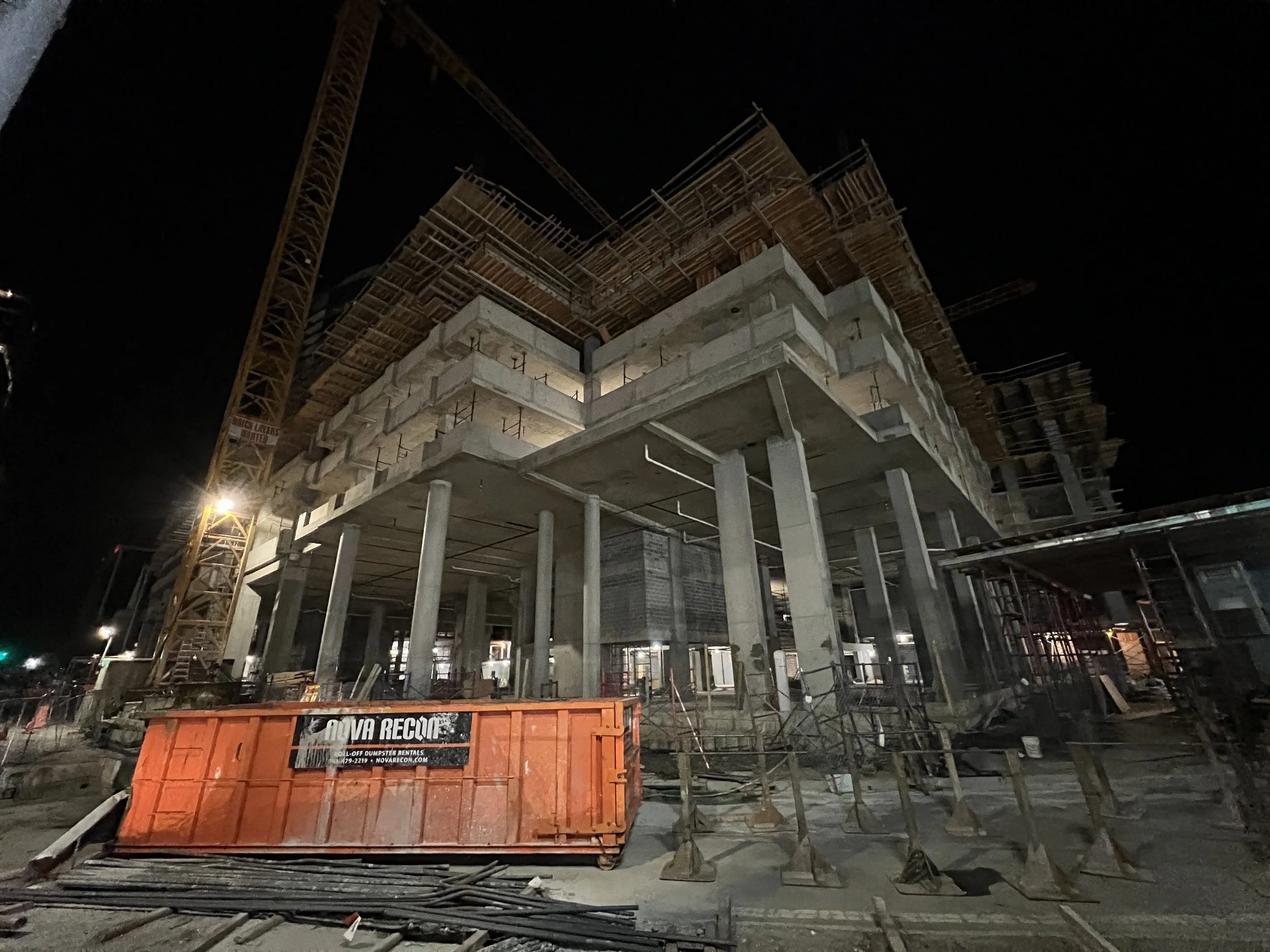 Nighttime view of a multi-story building under construction with scaffolding, cranes, and construction materials visible.
