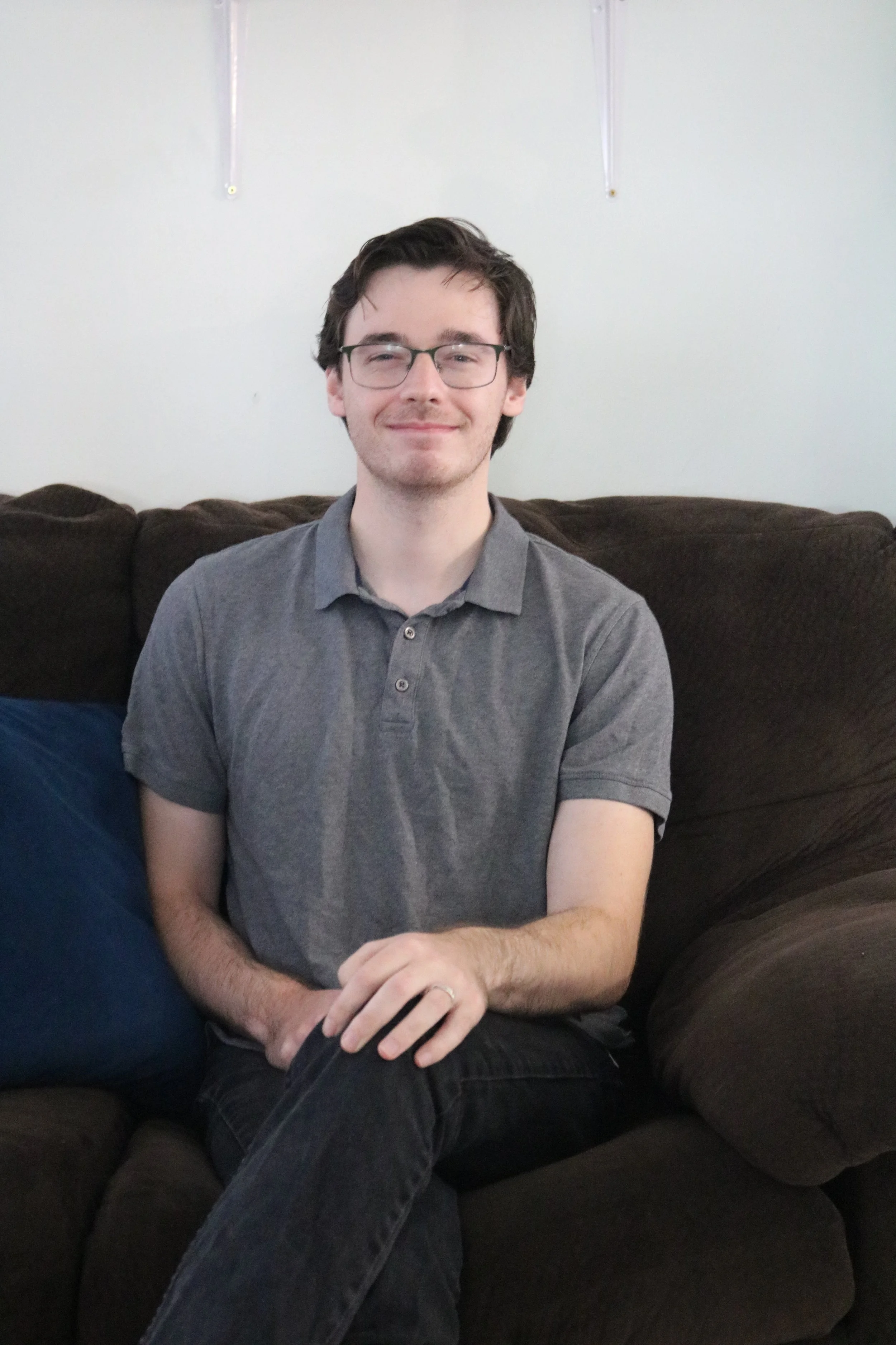 A young man with glasses wearing a gray polo shirt sitting on a brown couch.