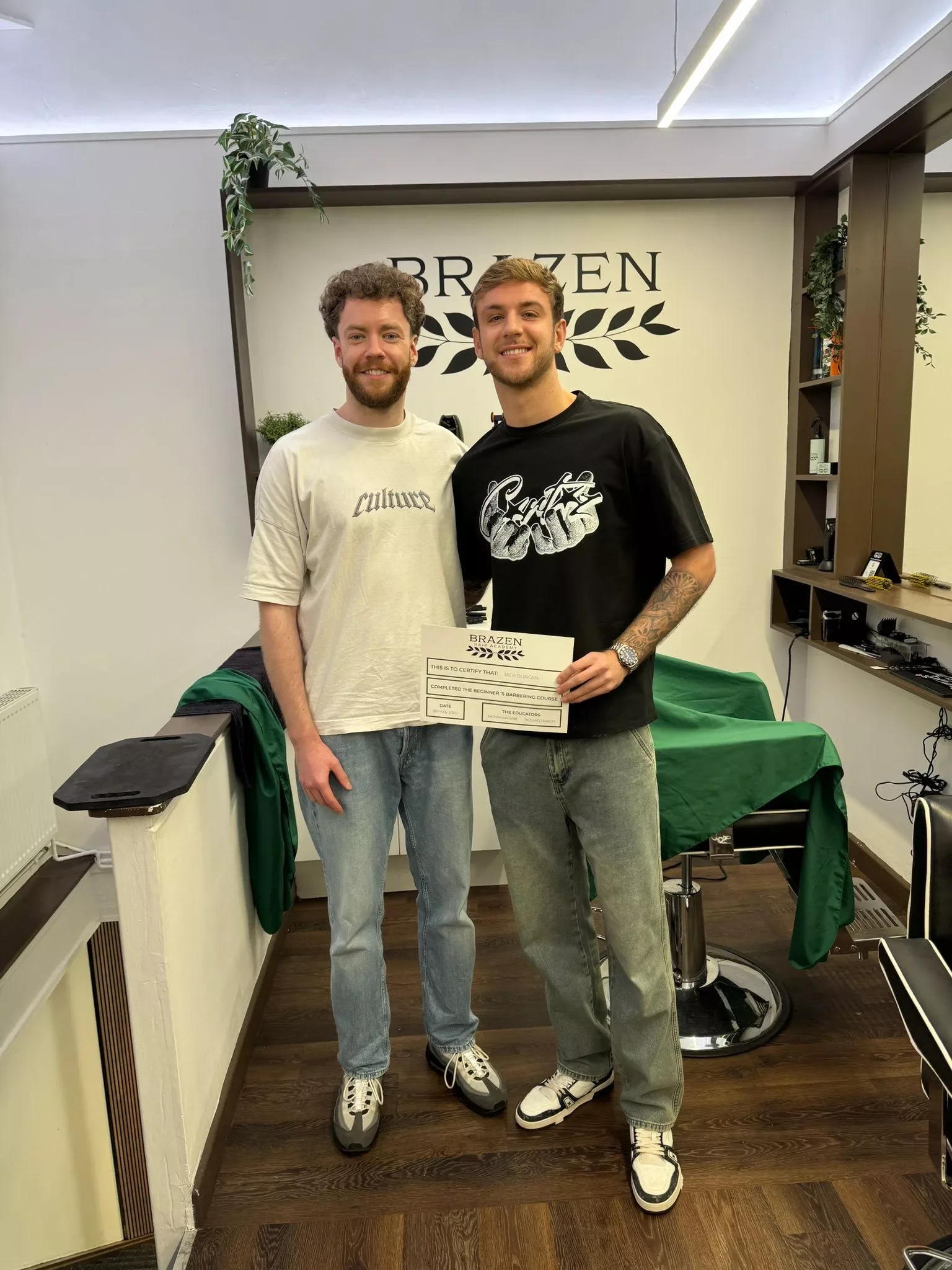 Two young men standing side by side inside a barbershop, smiling, with one holding a certificate. The barbershop has a sign that reads 'BRAZEN' on the wall, various hair products on shelves, and a green cloth-covered table. The men are dressed casually, wearing sneakers.