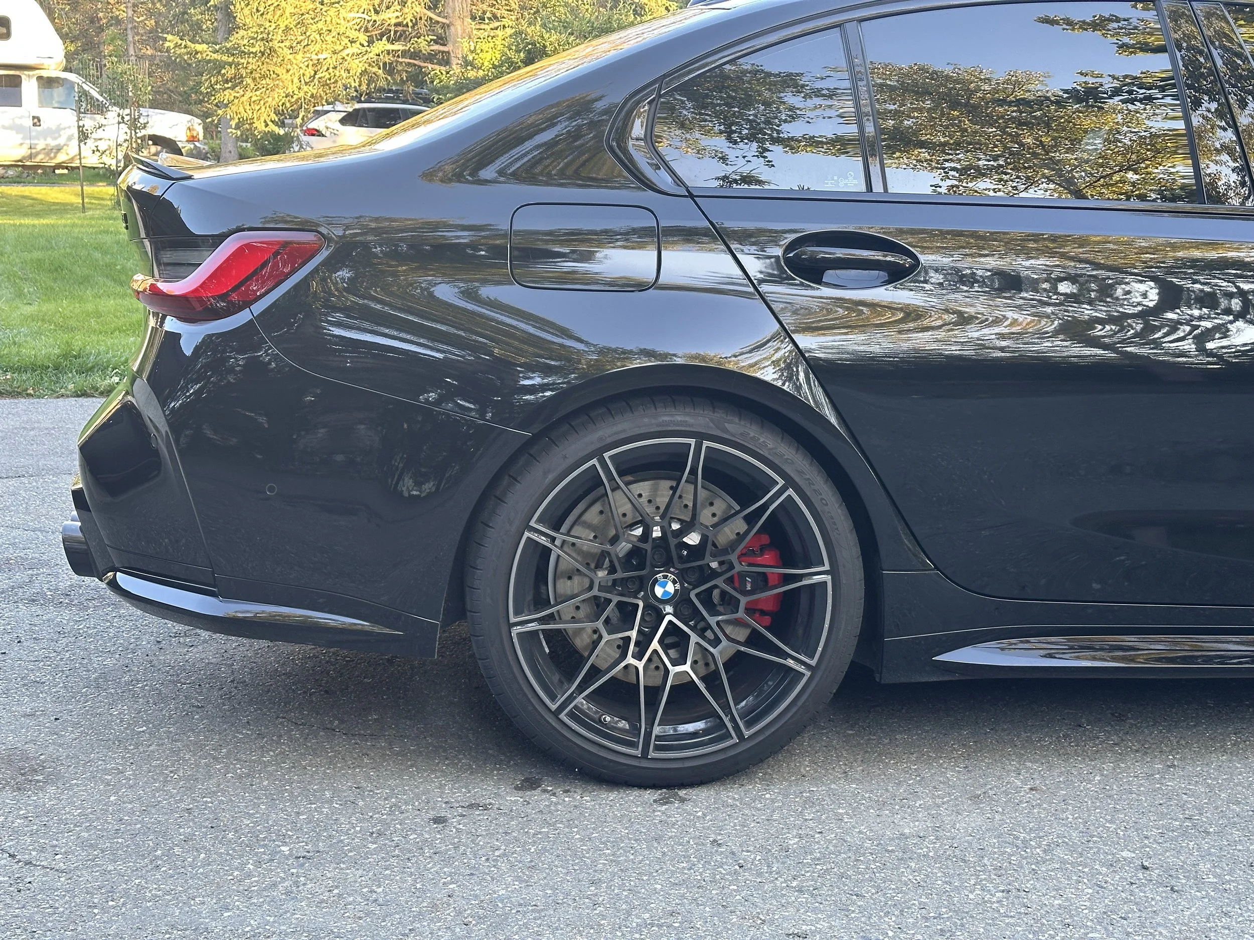 Close-up of the rear side of a black BMW M4 sports car, showing the wheel, red brake caliper, and part of the tail light, parked on asphalt with trees and other vehicles in the background.