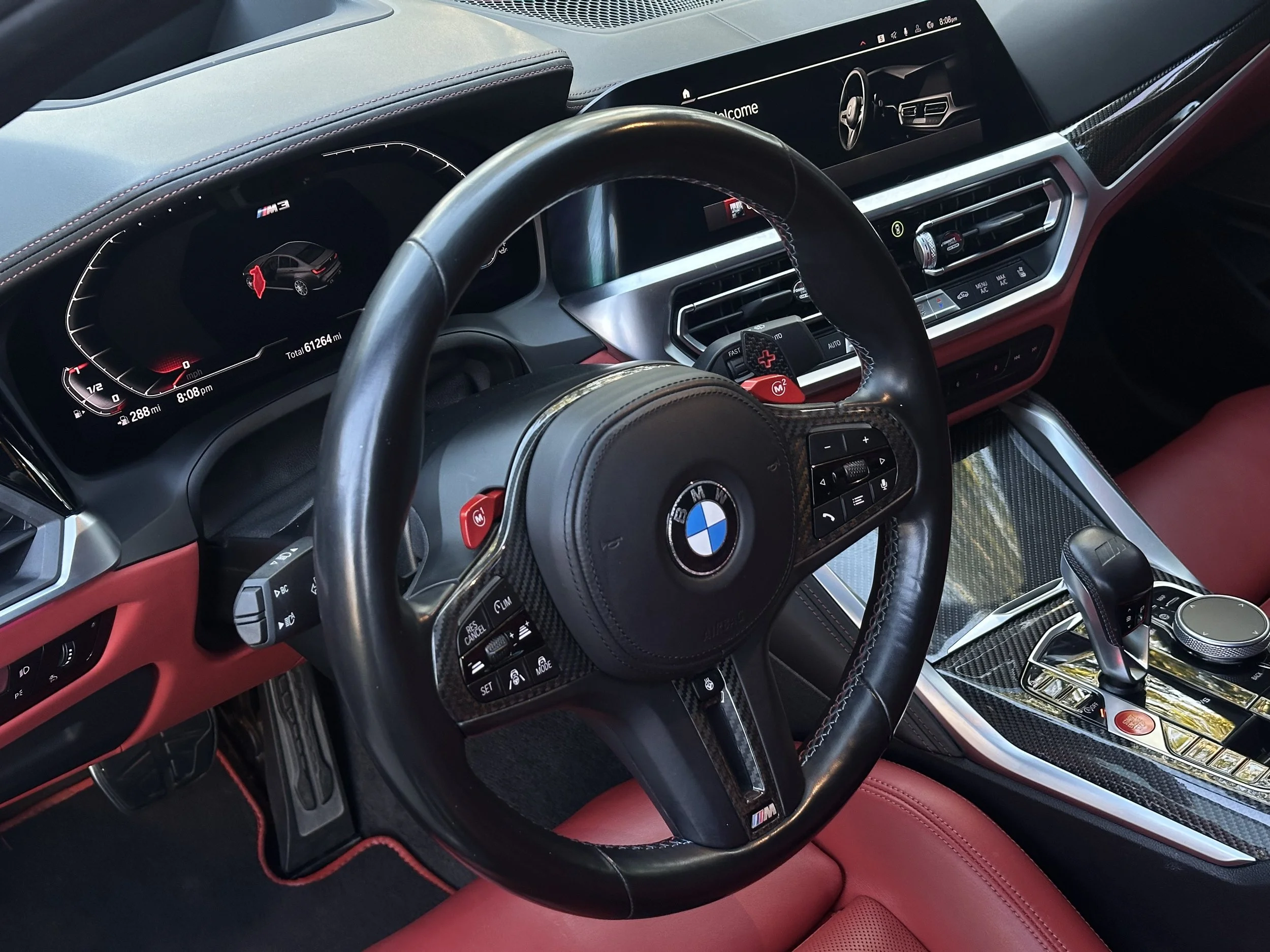 Interior view of a BMW M3's dashboard and steering wheel, featuring digital displays, carbon fiber accents, and red leather upholstery.