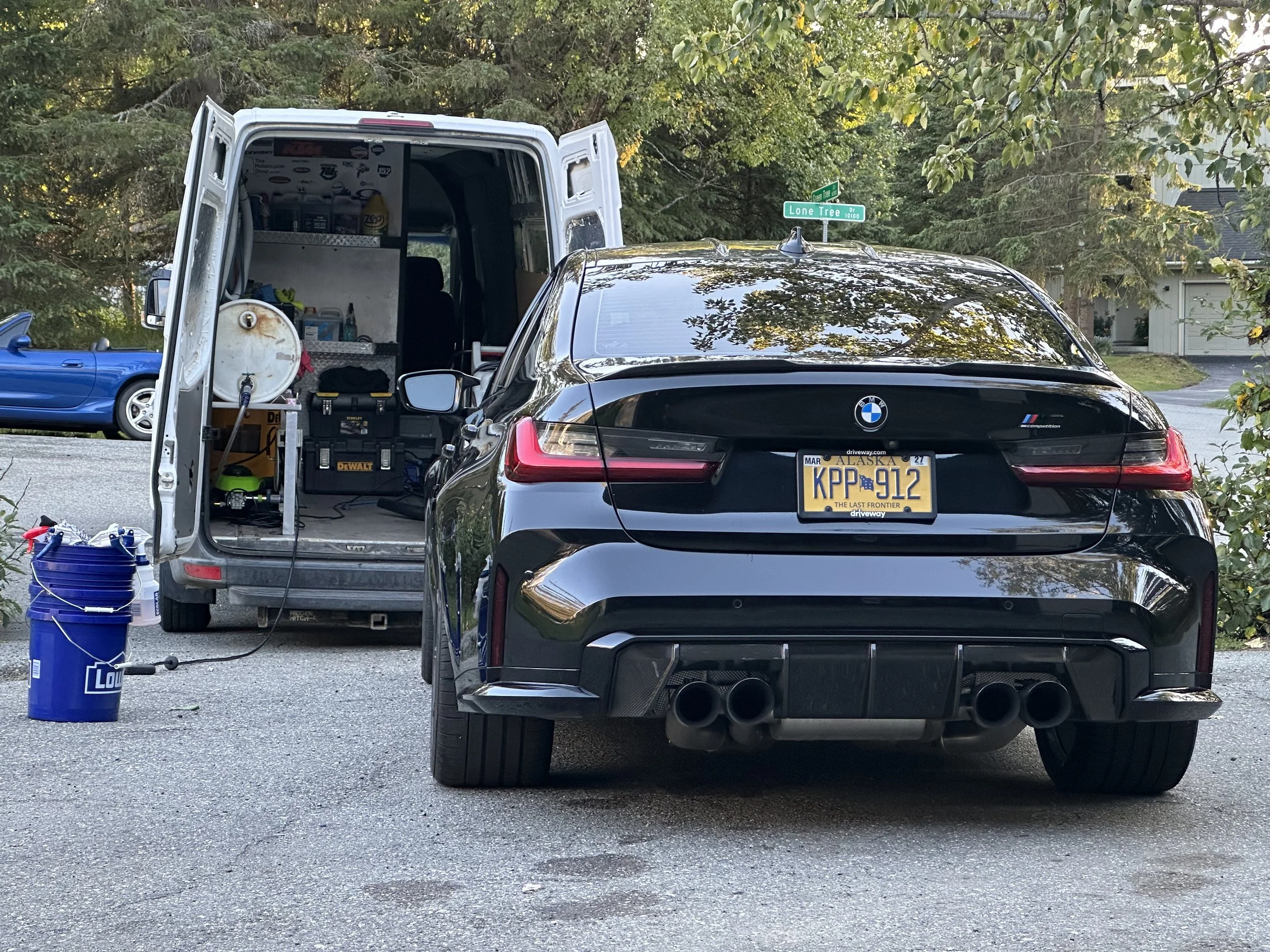 A black BMW parked in front of a white service van during daytime. The van has an open back with shelves containing tools and supplies. There is a blue bucket with a hose connected to a water pressure washer on the ground in front of the van. A stree
