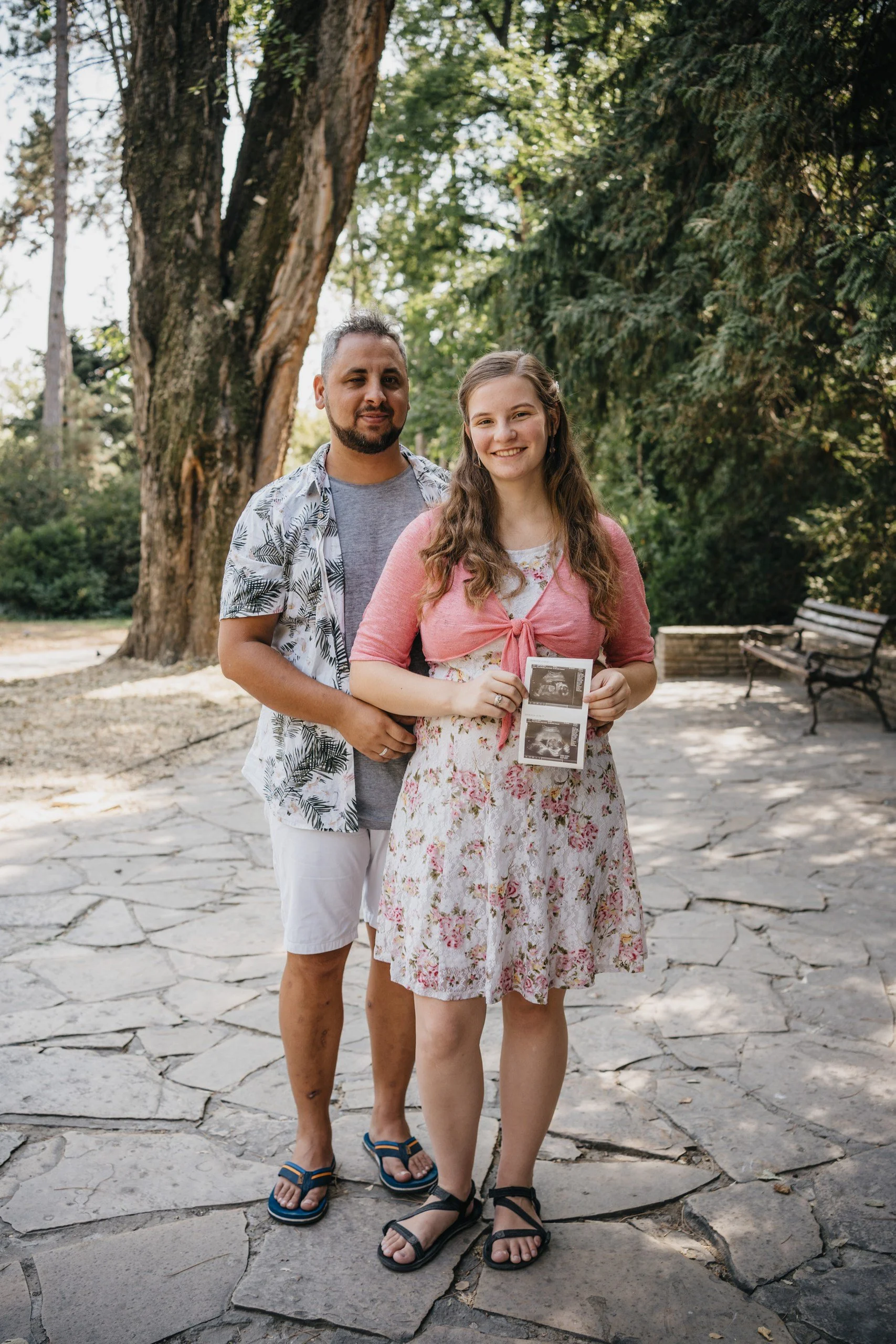 A young woman holding ultrasound images stands outdoors with a man behind her, both smiling. They are in a park with large trees and park benches.