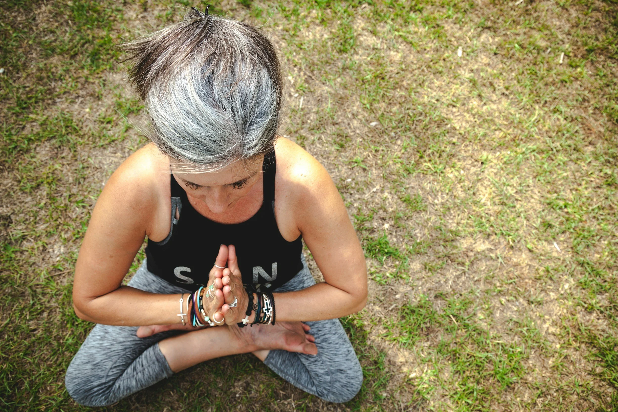 A woman with gray hair practicing yoga outdoors on a grassy field is sitting cross-legged with her hands in a prayer position.