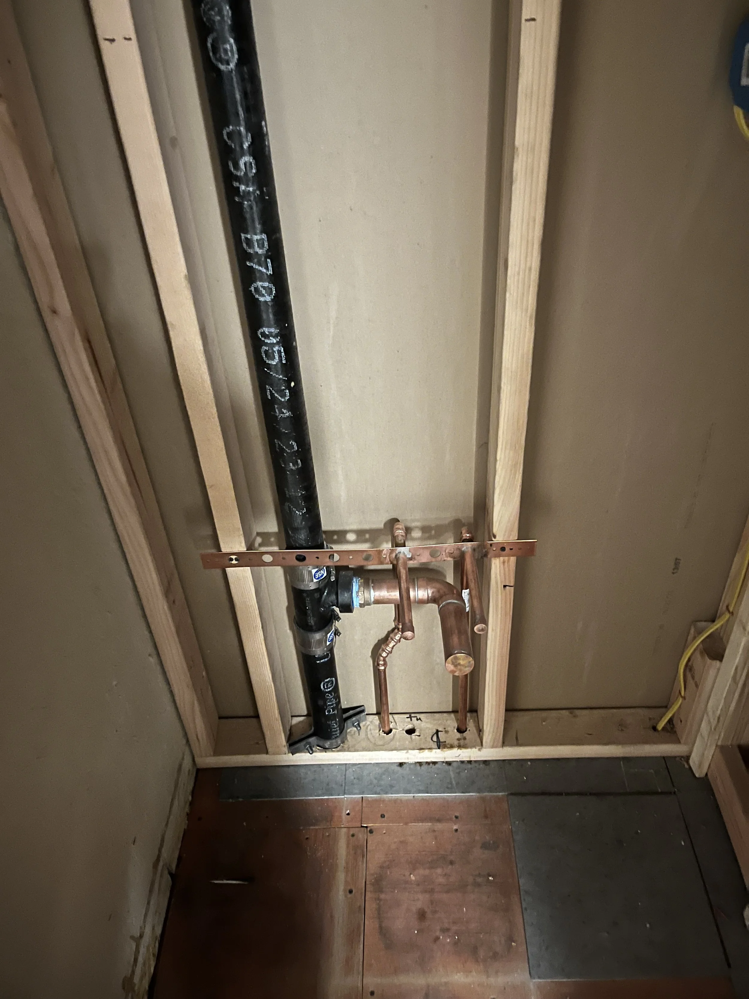 Plumbing pipes and fixtures in a wall with wooden framing, including a black pipe, copper pipes, and a copper tee fitting, with wiring visible on the right side.