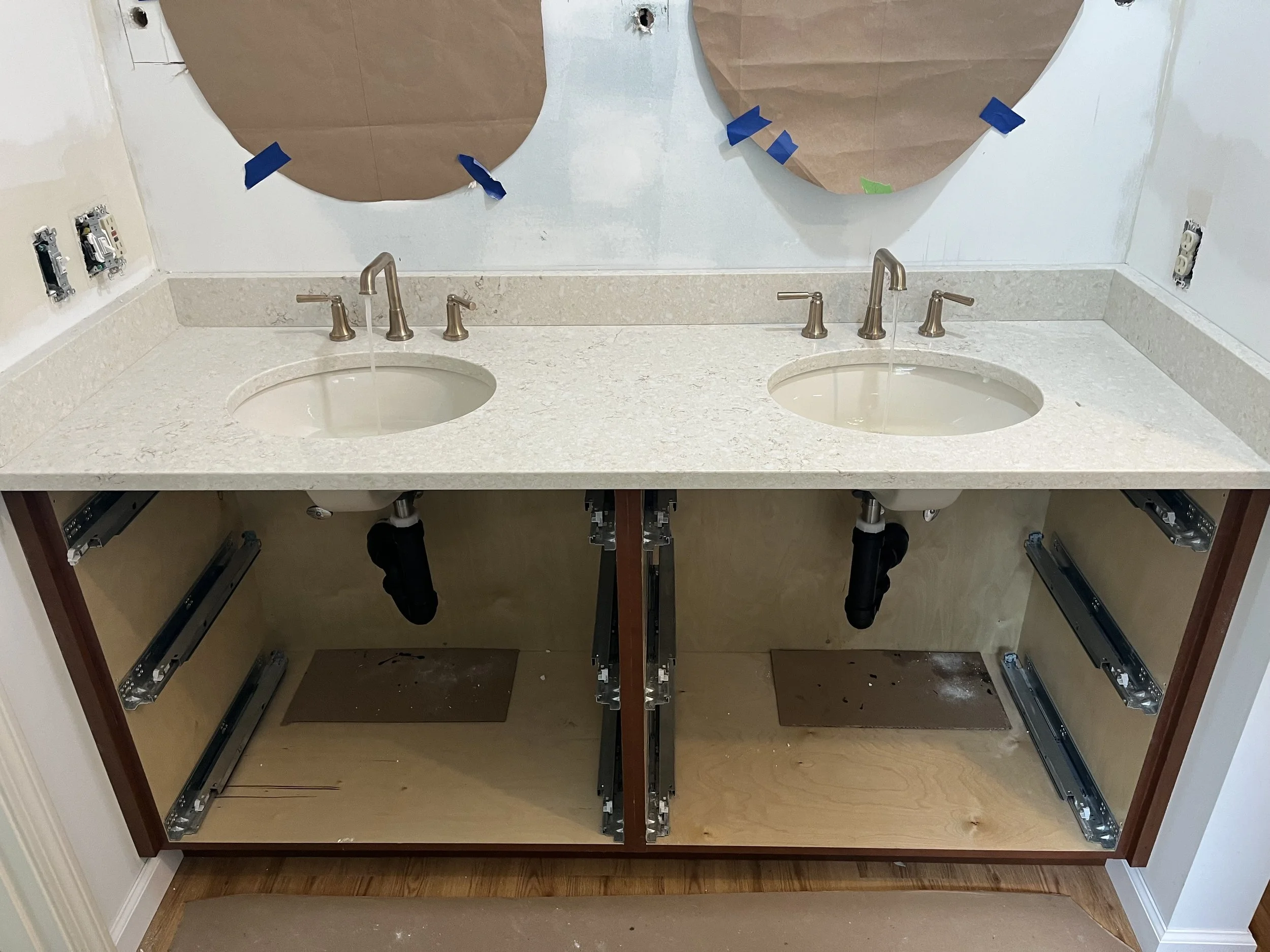 A bathroom vanity with two sinks and faucets, under construction, with no cabinet doors and exposed plumbing and drawers. Two mirrors covered with brown paper and blue painter's tape are above the sinks.