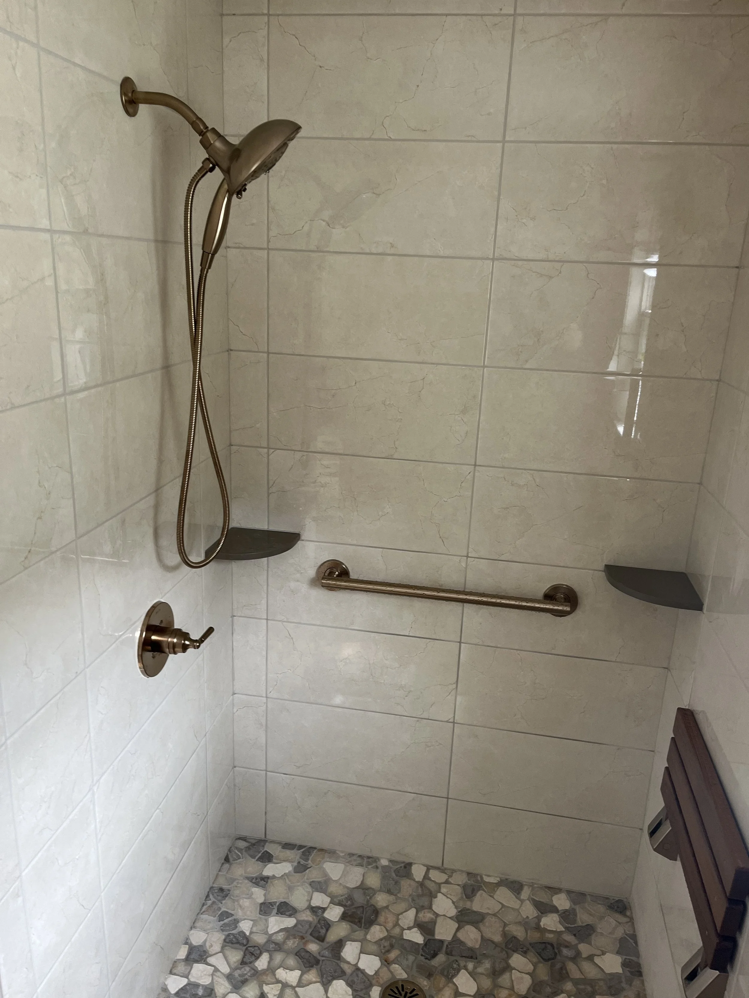 Walk-in shower with beige tiled walls, a handheld showerhead, a grab bar, two corner shelves, a wooden bench, and a pebble stone floor.