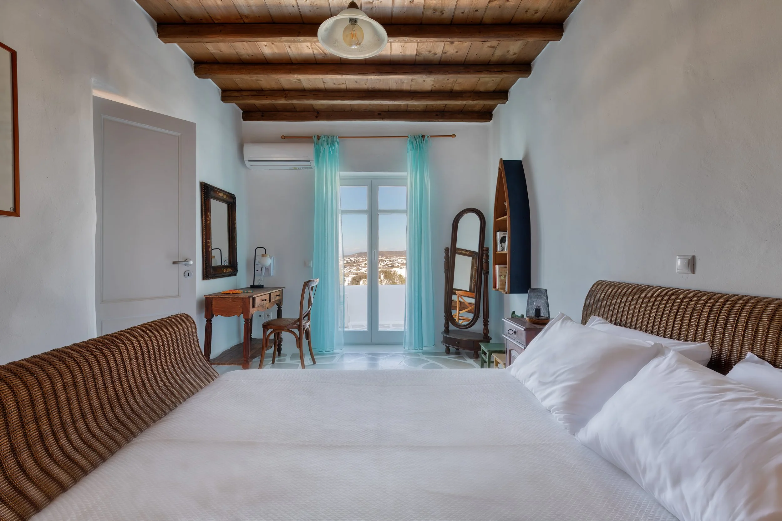 Bright double bedroom with wood-beam ceiling, A/C, desk, and balcony doors with panoramic views, Villa Zanetti Mykonos.