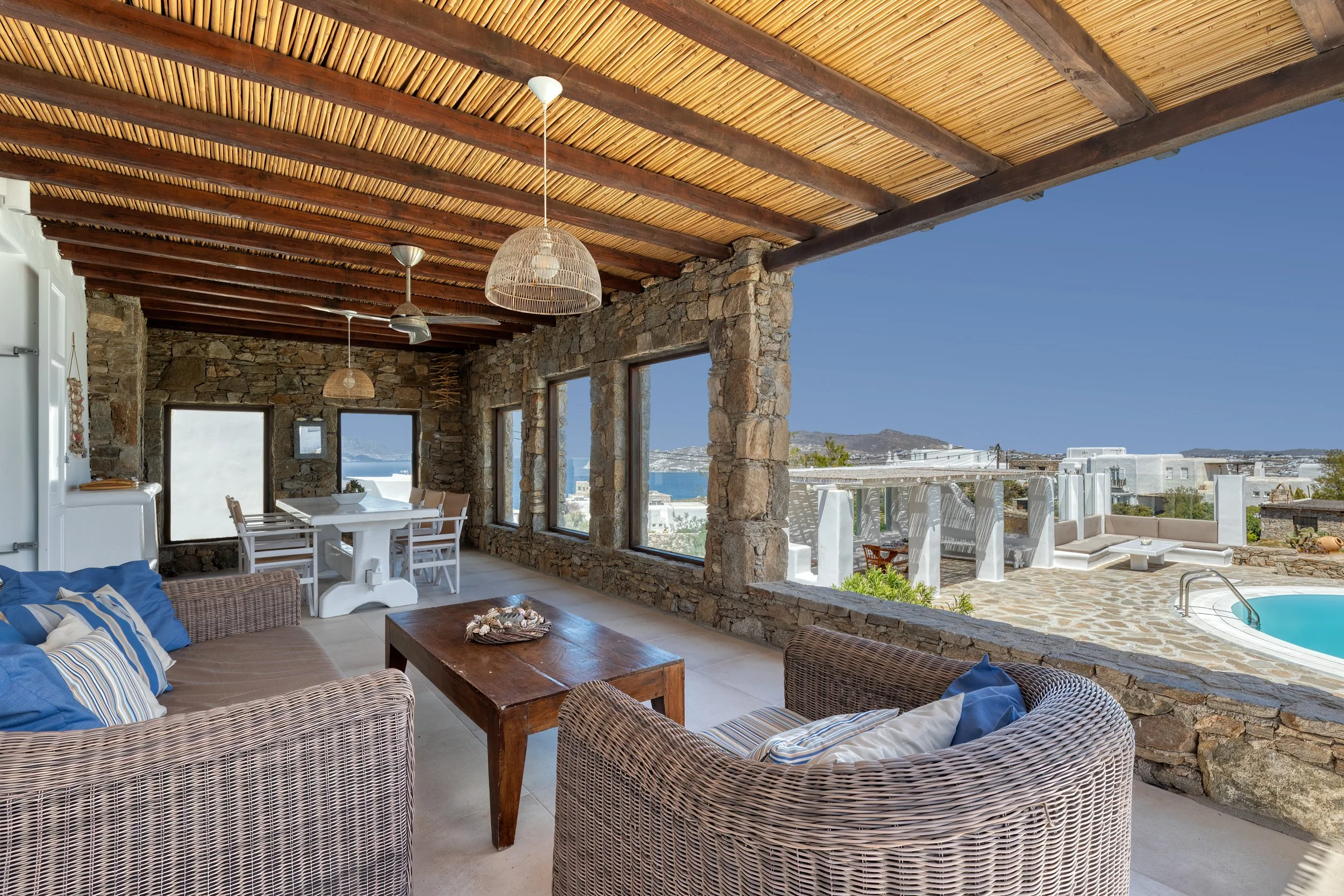 Outdoor living area under a shaded pergola with lounge seating and dining areas. Views of the pool, sea, and Mykonos.