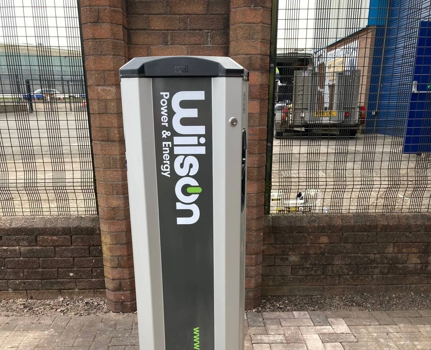 Electric vehicle charging station branded WiSUS, positioned outdoors near a brick wall and a metal fence, with vehicles and storage units in the background.