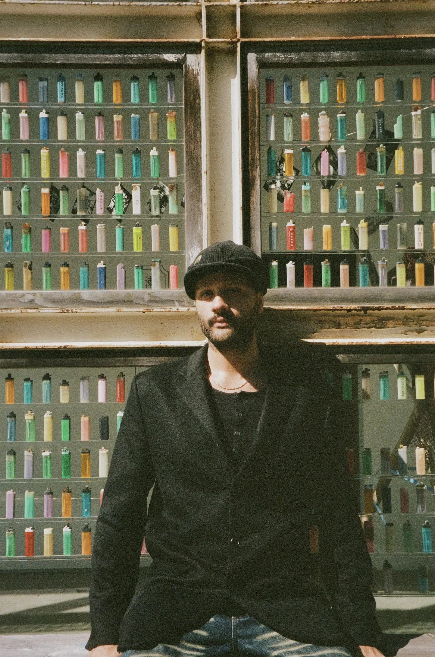 A man in a black coat and hat sitting in front of a display of colorful items, possibly lighters, in a rustic setting.