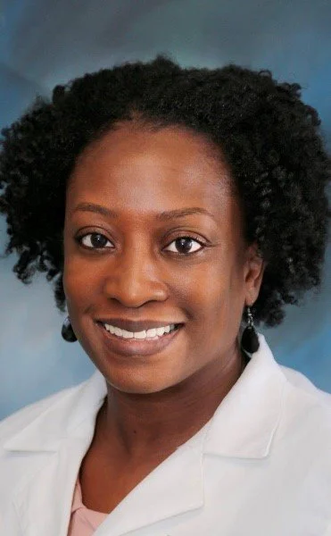 A woman with natural curly hair smiling, wearing a white coat and earrings, against a blue background.