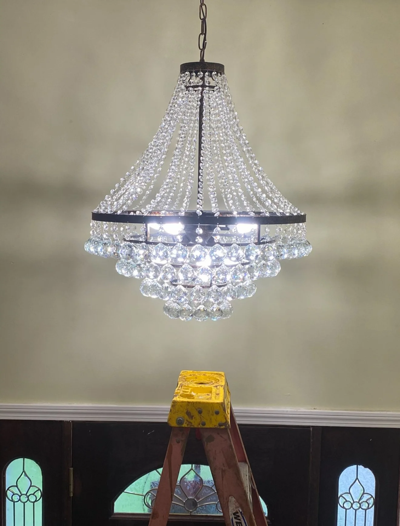 A chandelier with crystal beads hanging from a black metal frame, illuminated by several light bulbs, and a yellow step ladder underneath in a room with green walls.