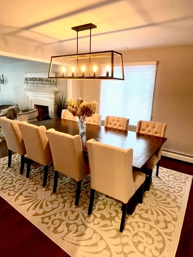 Dining room with a wooden table, ten beige upholstered chairs, a glass vase with flowers, a modern chandelier, a window with blinds, and a patterned rug.