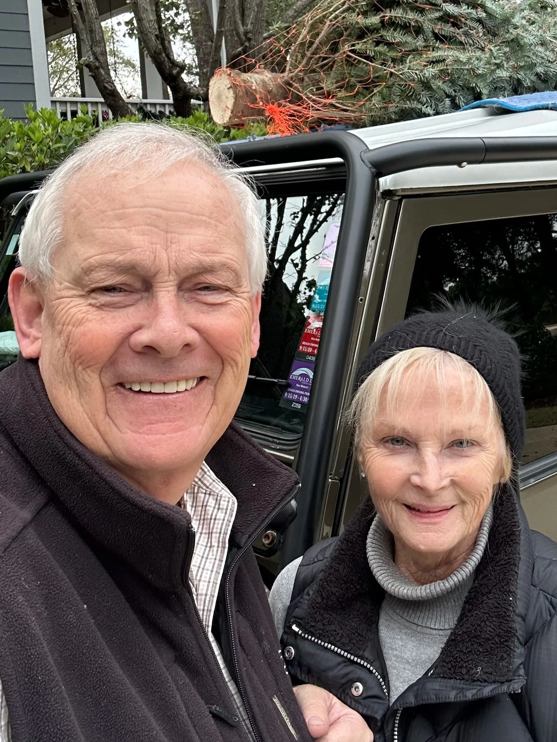 A smiling elderly man and woman taking a selfie outdoors in front of a vehicle with a large tree fallen on top of it.