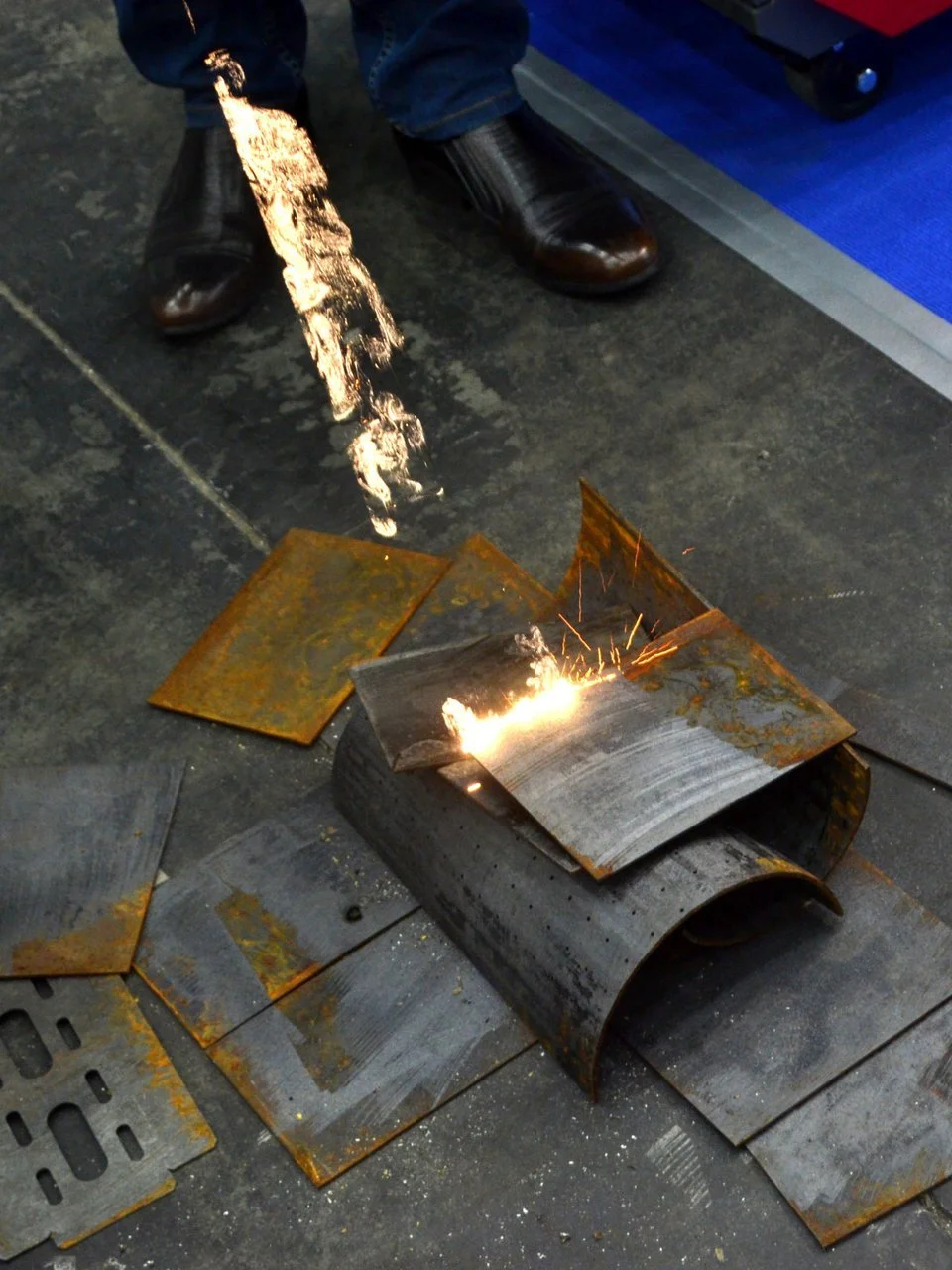 Person laser cleaning metal sheets, sparks flying from the laser cleaning and restoration process on a concrete floor.