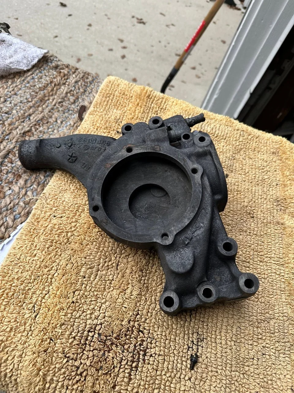 A black car part on a yellow towel, placed on a textured surface near an open garage door showing the difference after being laser cleaned.