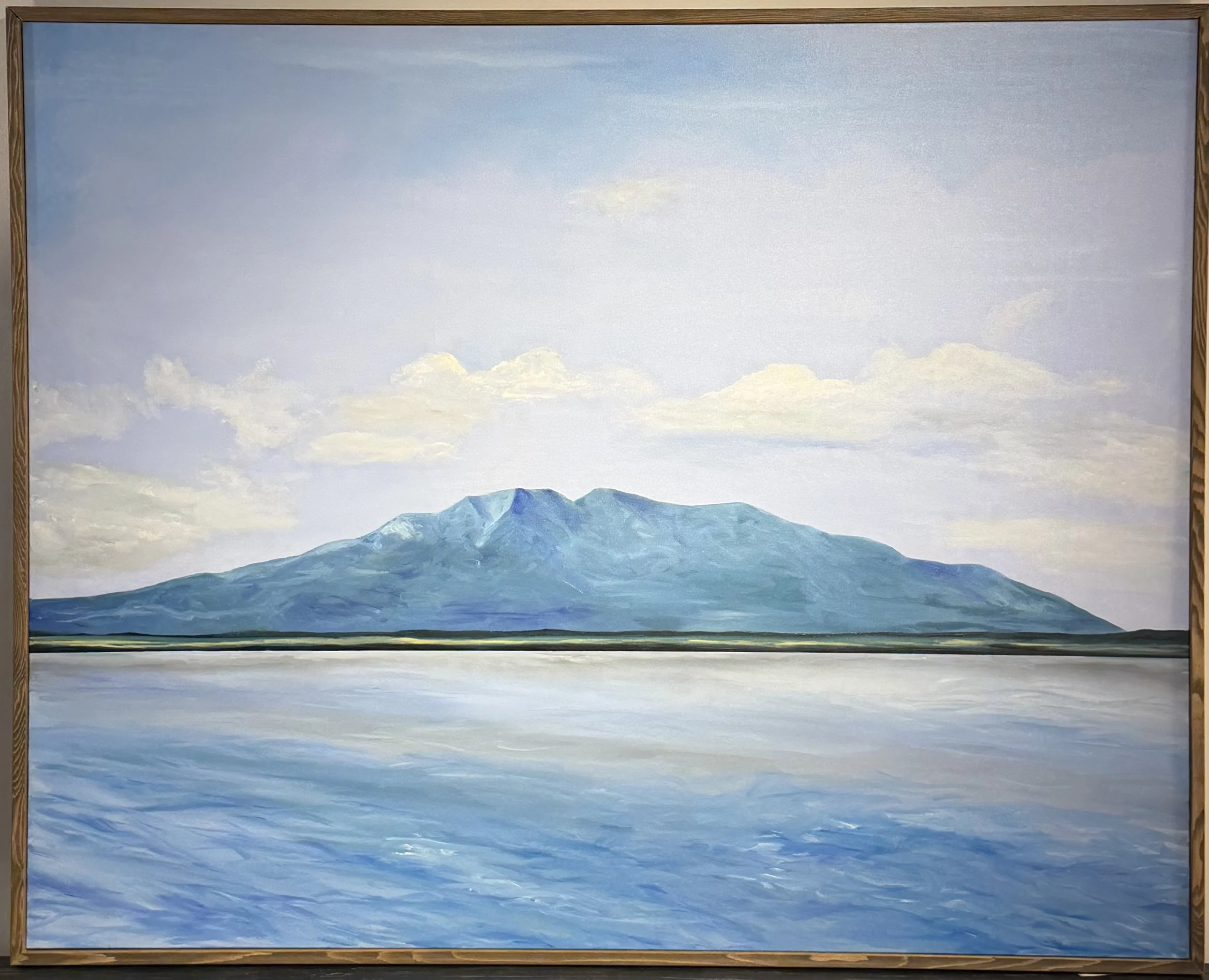 Large oil painting of a solitary blue-green mountain rising above a flate horizon, its reflection shimmering instill water below. Soft lavendera nd white clouds dift throuh a pale sky. Displayed in a natural wood float frame.
