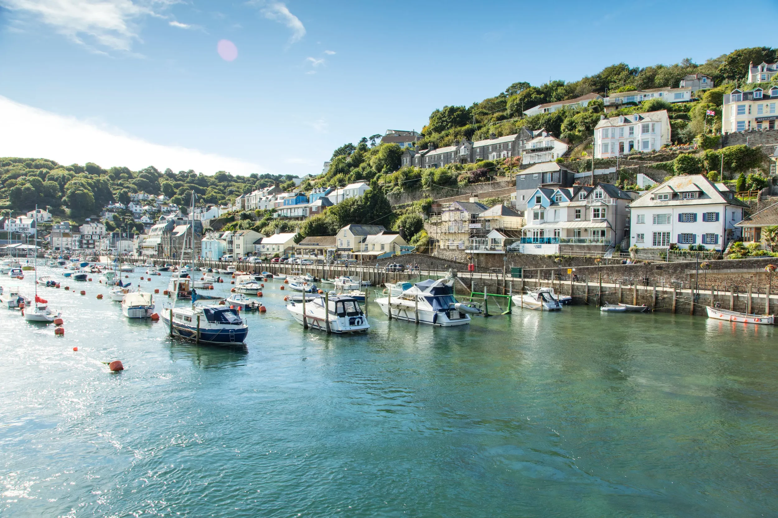 A harbor filled with boats and yachts docked along a pier, with a hillside in the background dotted with colorful houses, under a bright blue sky.