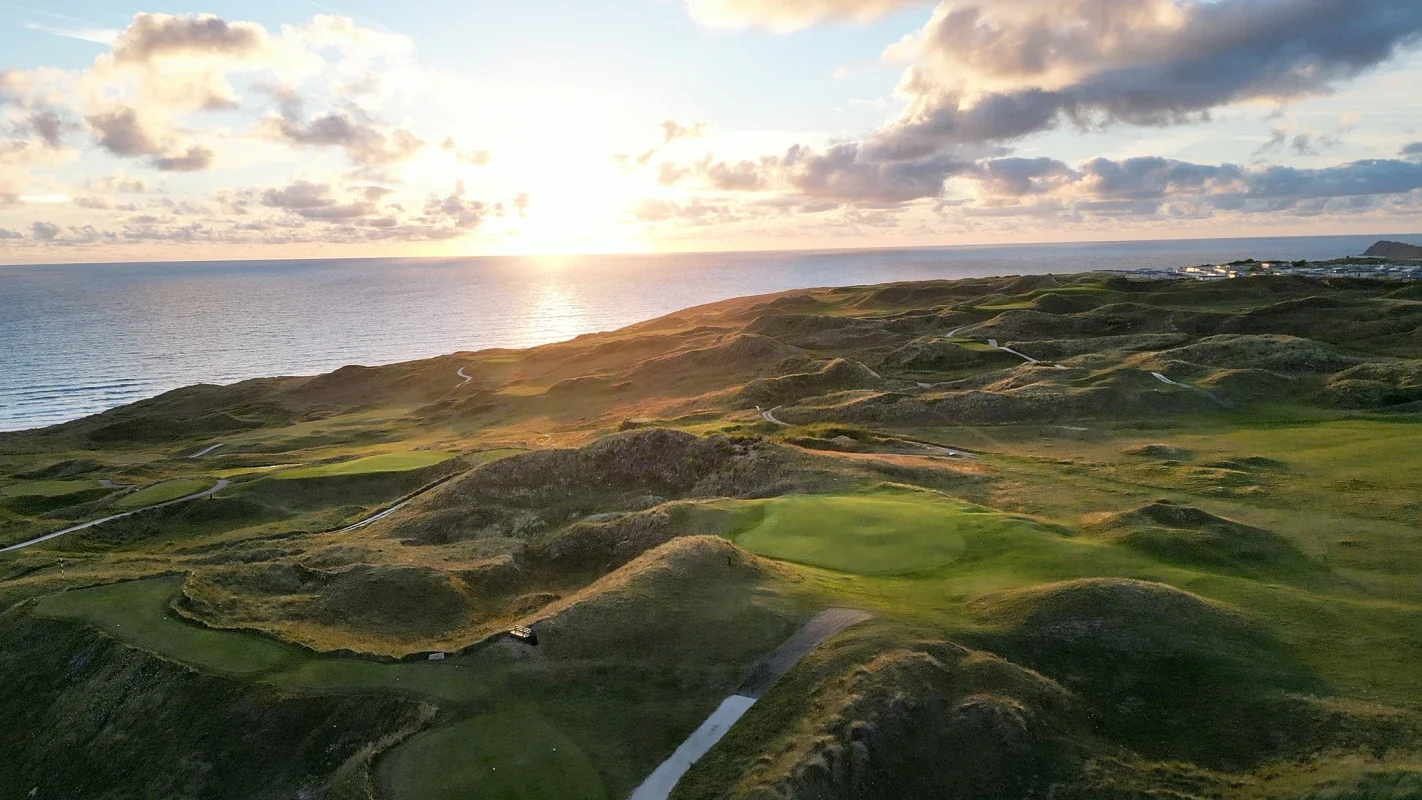Aerial view of a coastal golf course with green fairways and sand traps, overlooking the ocean with the sun setting in the sky, partly cloudy.