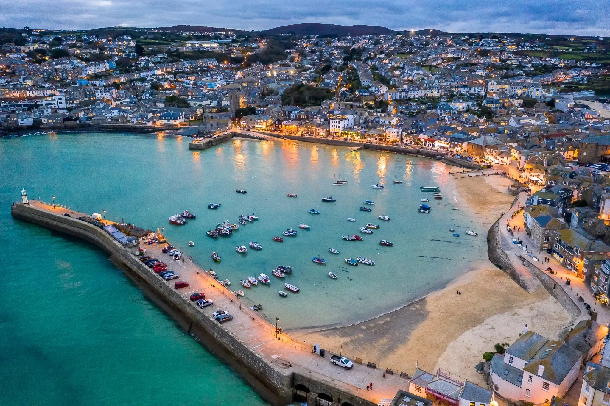 Aerial view of St Ives, in the UK. A coastal harbor with boats, a sandy beach, and a town with buildings along the shoreline during dusk.
