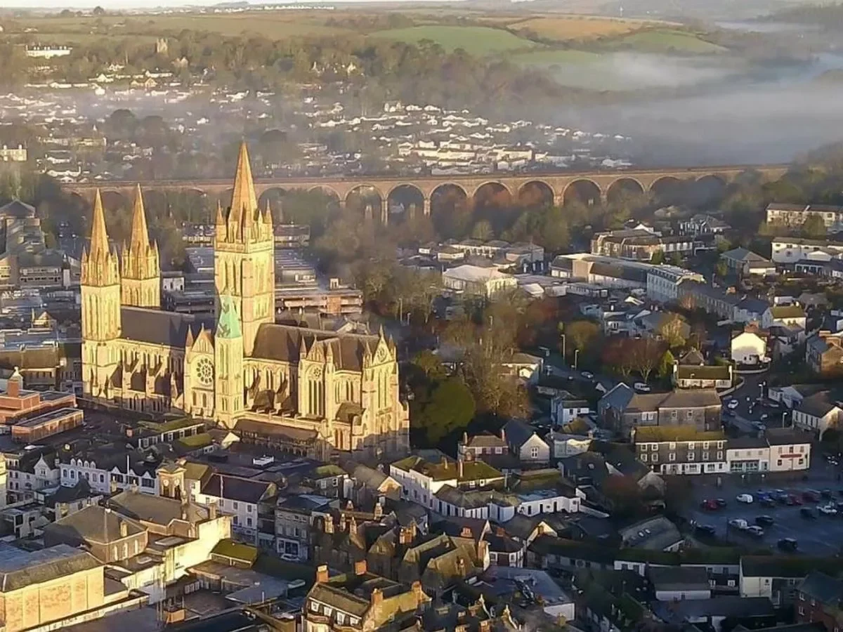 Aerial view of a historic city with a large Gothic-style church featuring multiple spires, residential and commercial buildings, a railway bridge, and a misty background with hills.