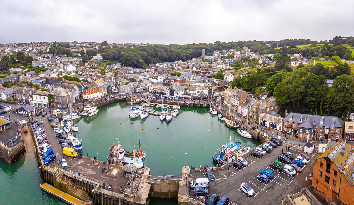 Padstow harbor with numerous boats docked, surrounded by a town with colorful buildings, a parking lot, and lush green hills in the background.