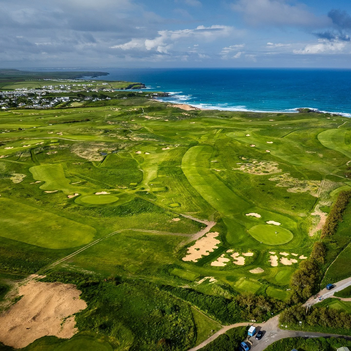 Aerial view of a coastal golf course with lush green fairways, sand bunkers, and a view of the ocean in the background under cloudy sky.