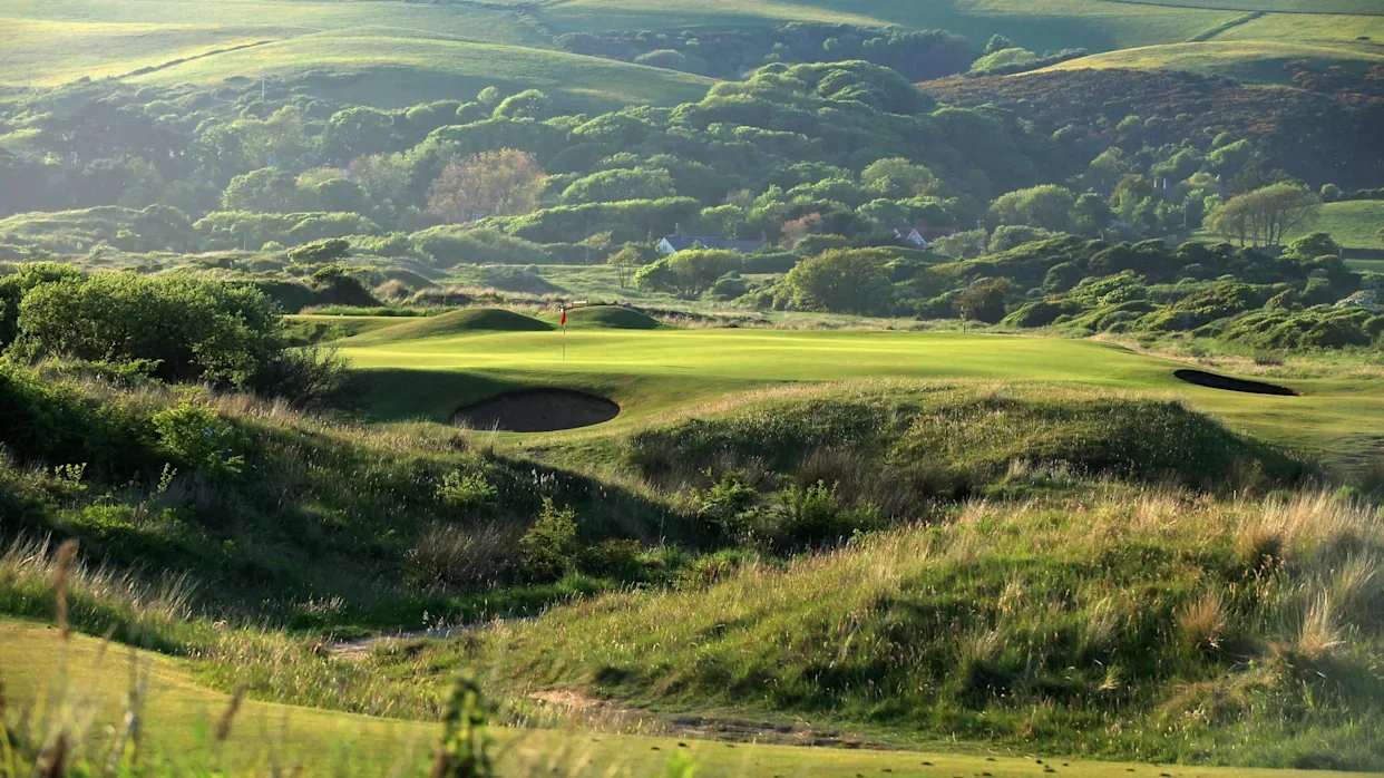 A scenic golf course green with a flag in the distance, surrounded by sand bunkers and lush grass, set against rolling hills and dense trees in the background.