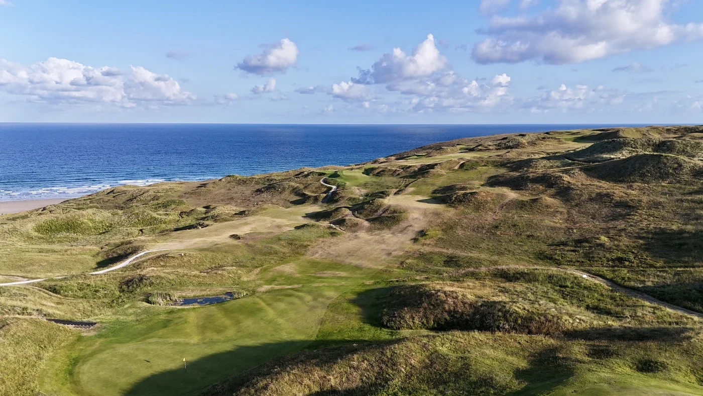 A coastal golf course with green fairways and sandy dunes overlooking the ocean under a partly cloudy sky.