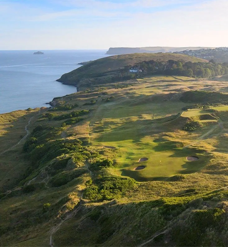 A coastal landscape featuring rolling green hills and a golf course with sand traps, overlooking the ocean with a few small islands and a distant shoreline in the background.