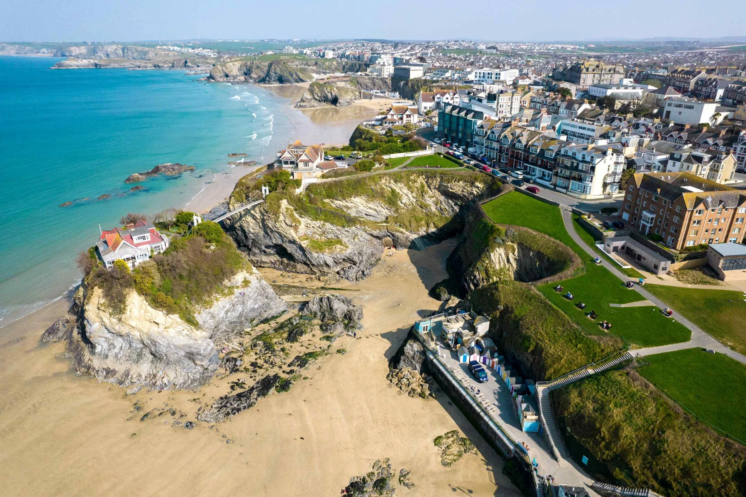 Aerial view of a coastal town with a sandy beach, cliffs, and colorful buildings along the shoreline. Houses and apartments are visible on the hillside, with a park area featuring benches and pathways. The ocean is turquoise, and there are rocky outcroppings in the water.
