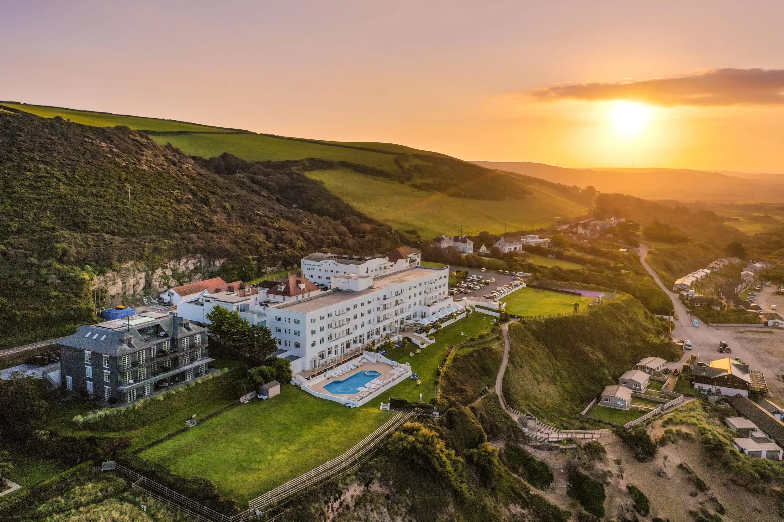 Saunton Sands Hotel. An aerial view of a hotel complex with a swimming pool, set on a hillside during sunset.