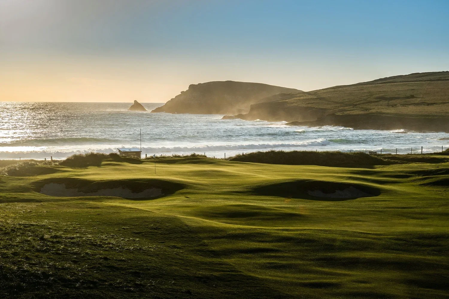 Golf course with sand traps near the ocean during sunset, with cliffs in the background and waves crashing against the shore.