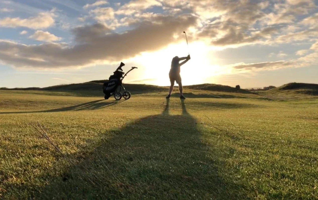 A person playing golf on a grassy course with the sun setting behind them, casting long shadows, and a golf bag on a trolley nearby.