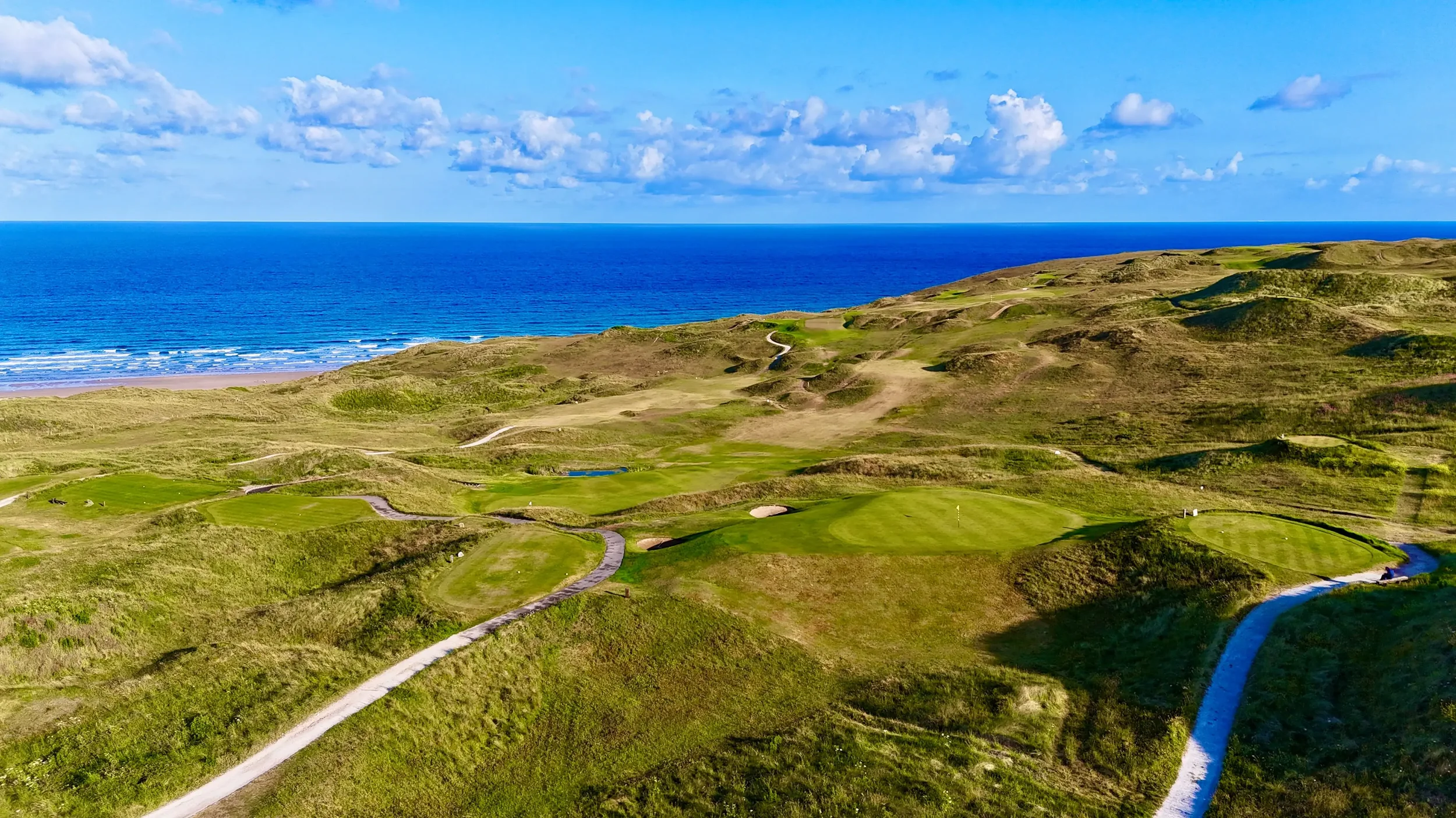 A scenic view of a golf course with green fairways and sand traps near the coast, with the ocean and sky in the background.