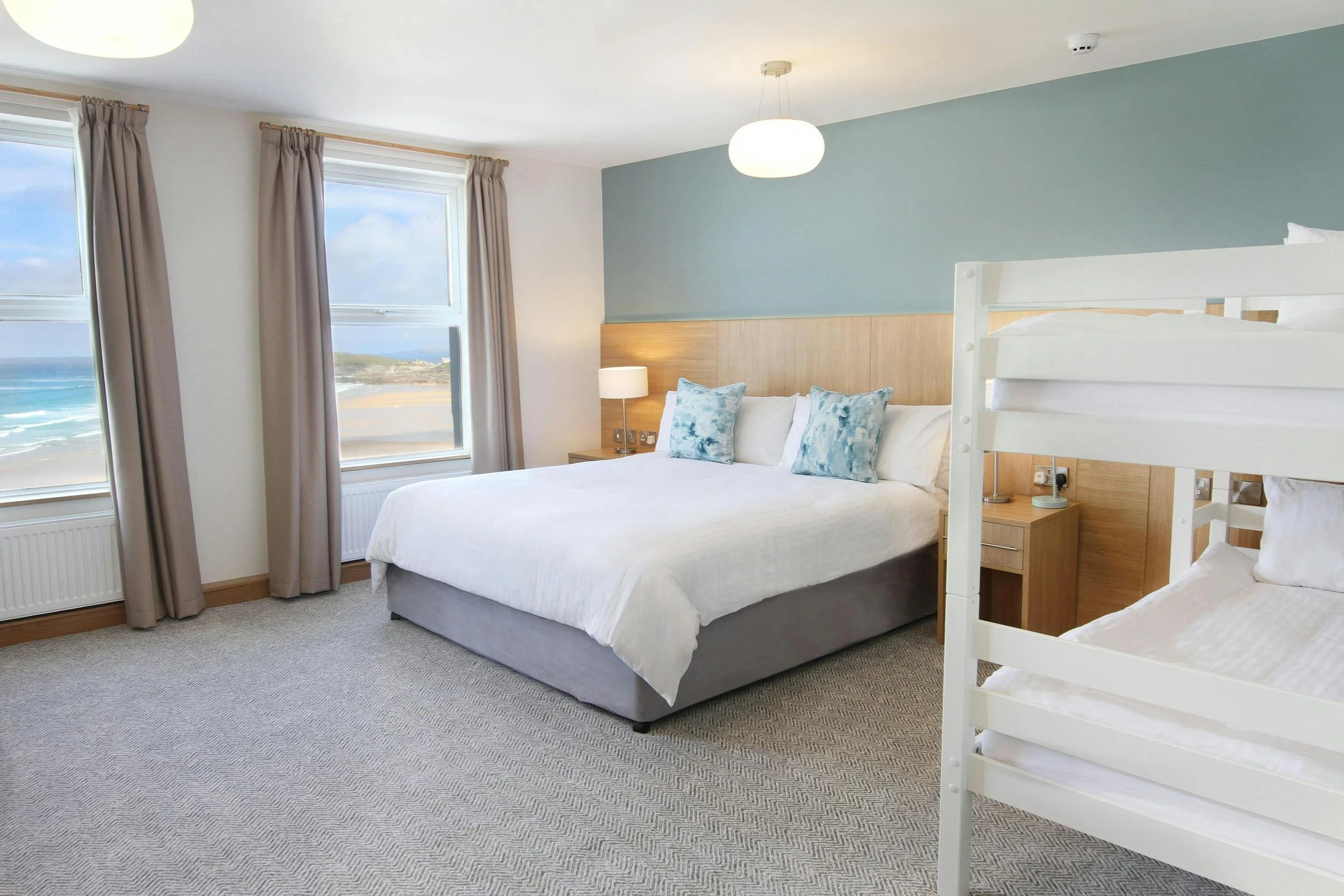 A hotel room with a large bed featuring four pillows, two decorative ones, and a bunk bed to the right. The room has two windows with beige curtains showing a beach, ocean, and sky outside. It is decorated with a wood headboard, nightstands, and lamps.