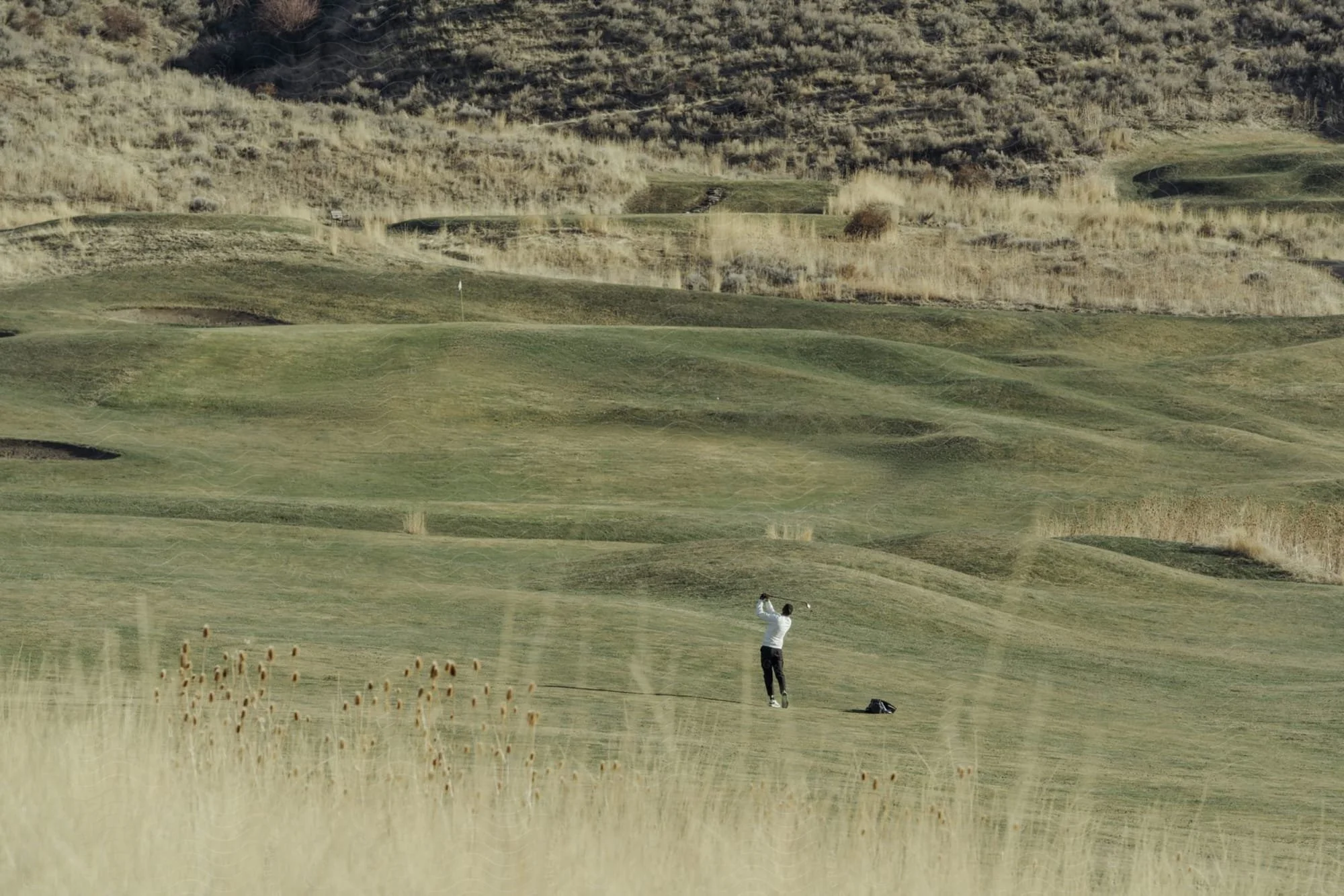 A person playing golf on a rolling, grassy golf course with hills and sparse vegetation in the background.