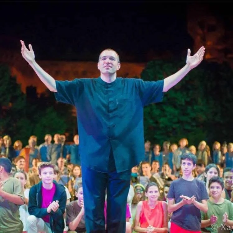 Gerard Pastor with his arms raised, receiving applause from the audience with children and musicians behind him on stage.