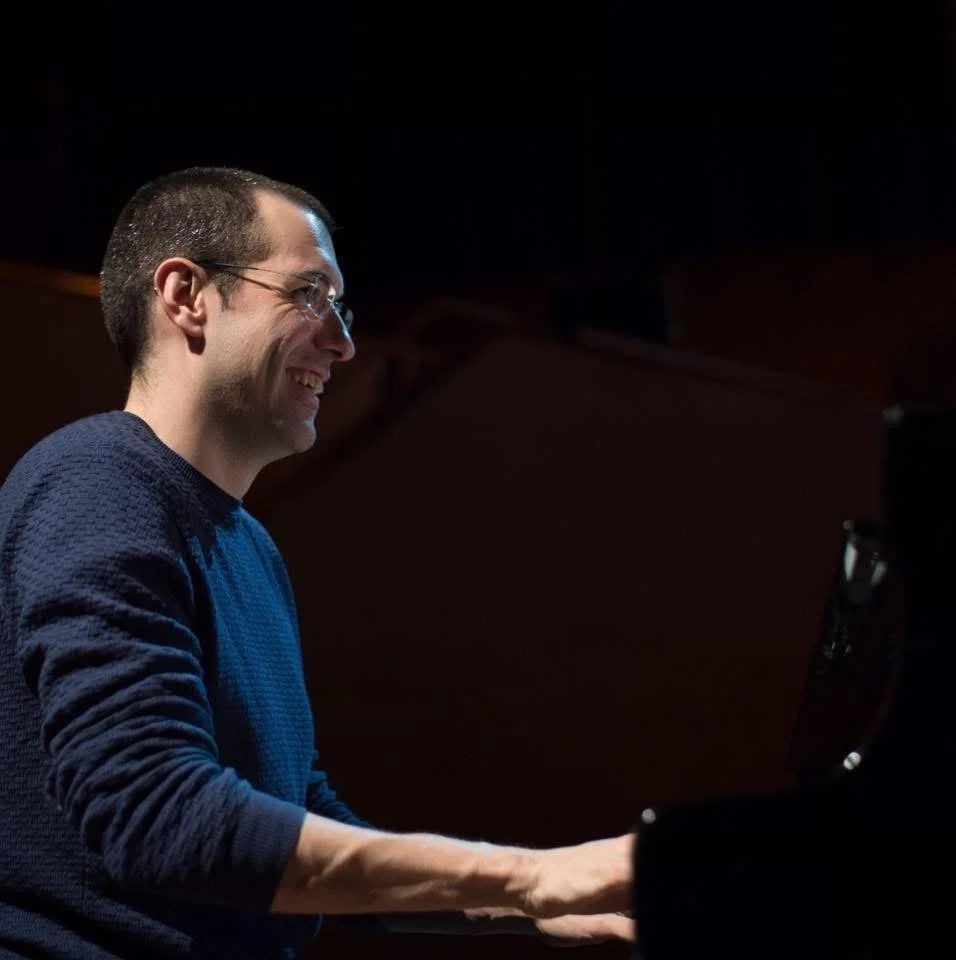 Side photograph of Gerard Pastor playing the piano during a rehearsal, wearing a blue sweater, with a completely black background. He smiles while looking forward.