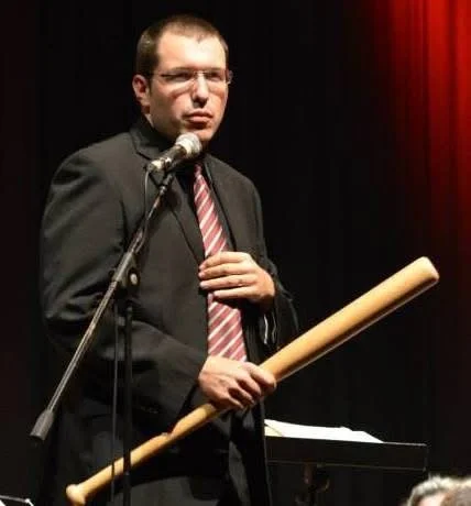 Gerard Pastor in a suit, speaking into a microphone and humorously holding a baseball bat.