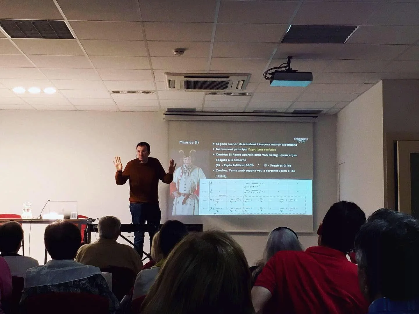 Photograph from the audience of a talk by Gerard Pastor. He wears a wireless microphone and raises his hands to illustrate a point; a musical score is projected on the screen.