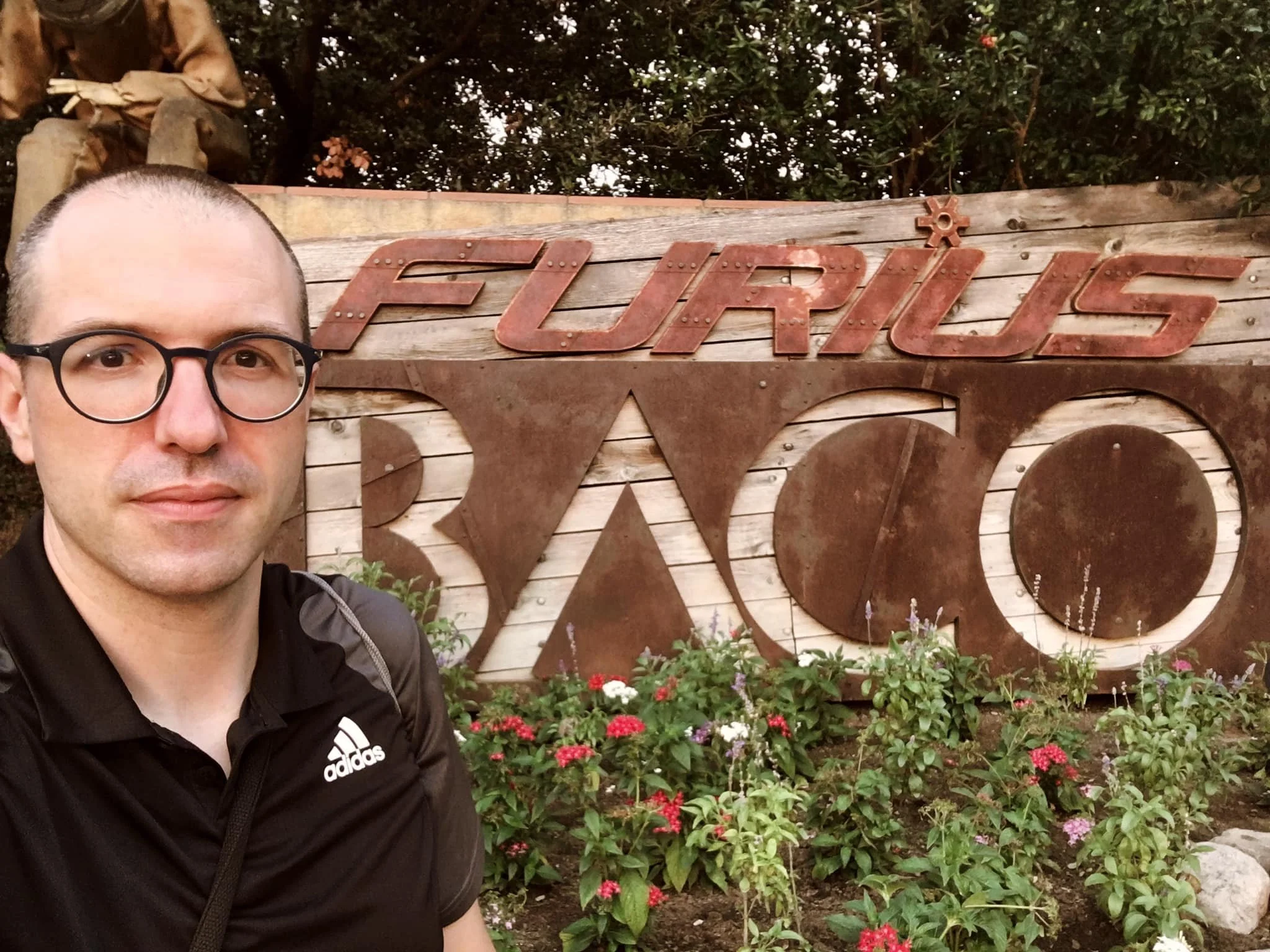 Gerard Pastor in a black Adidas sports T-shirt, with short hair and glasses in the foreground, in front of a metal sign reading ‘Furius Baco’ surrounded by flowers and plants. Entrance to the PortAventura attraction.
