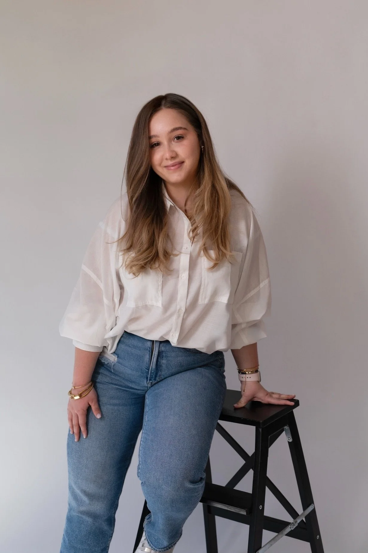 A young woman with long brown hair wearing a white blouse and blue jeans, sitting on a black stool against a plain light gray wall, looking at the camera with a slight smile.