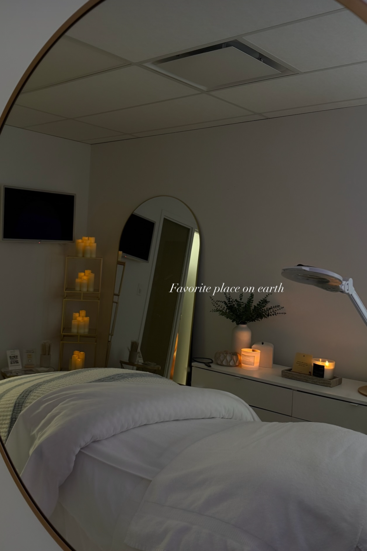 A cozy bedroom with a neatly made bed, soft lighting candles on a side table and shelves, a round mirror on the wall, a flat-screen TV, and a lamp with a plant on a dresser. Text overlay reads 'Favorite place on earth'.