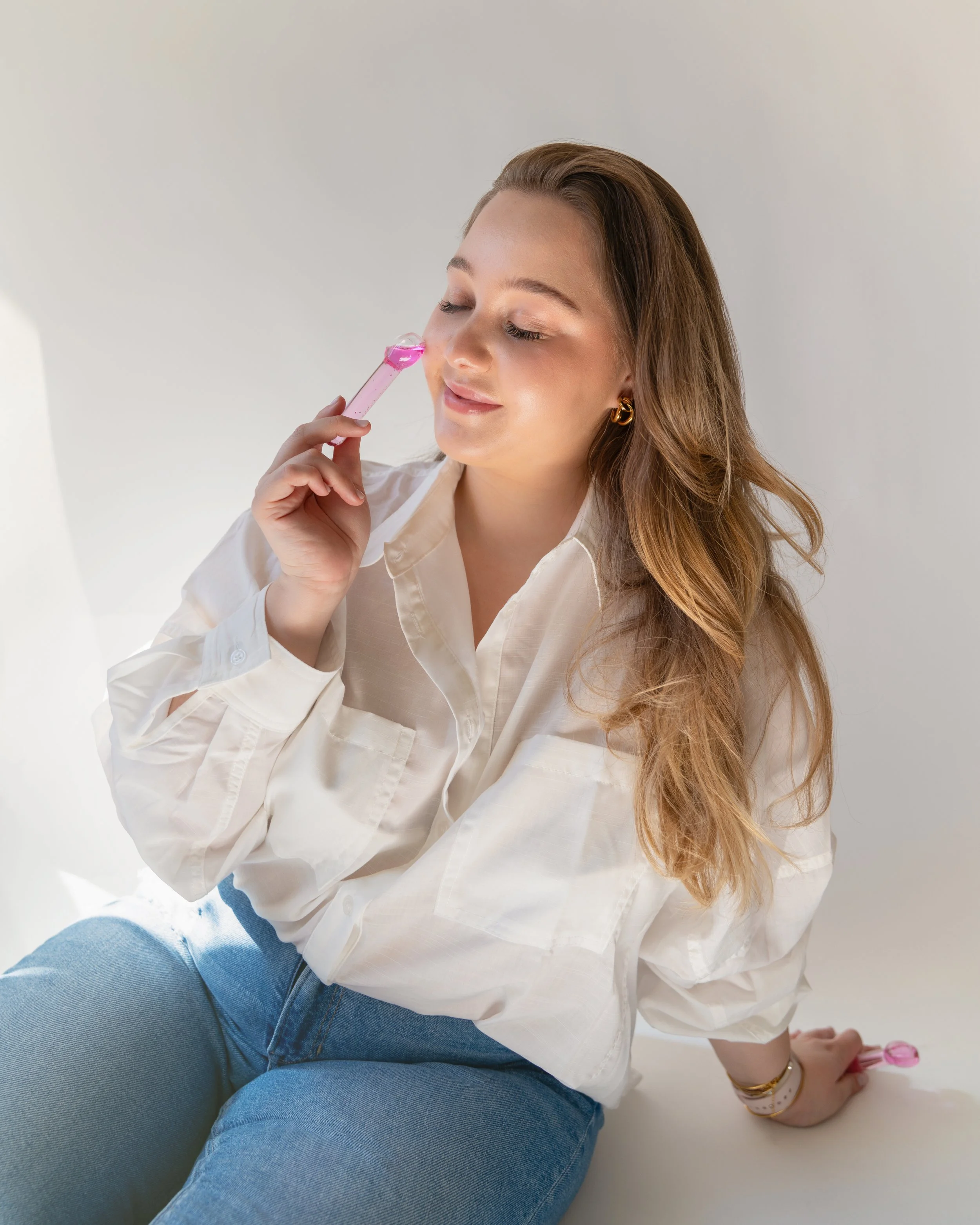 A woman with long wavy hair wearing a white shirt and blue jeans is sitting on a white surface, holding a pink bubble wand to her nose with her eyes closed.