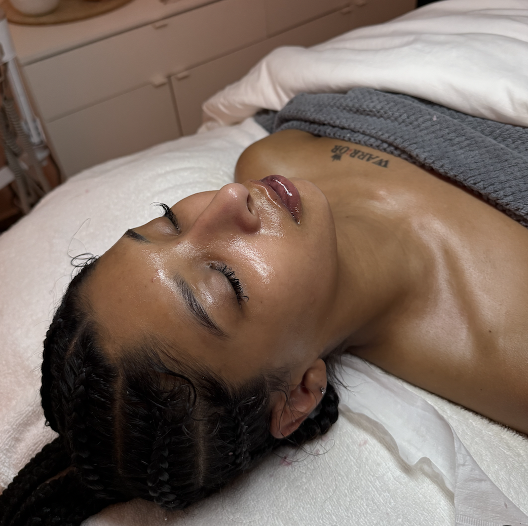 A woman with braided hair lying on a bed with her eyes closed, covered partially with a gray towel, in a room with beige furniture, appearing relaxed and possibly receiving a spa or skincare treatment.