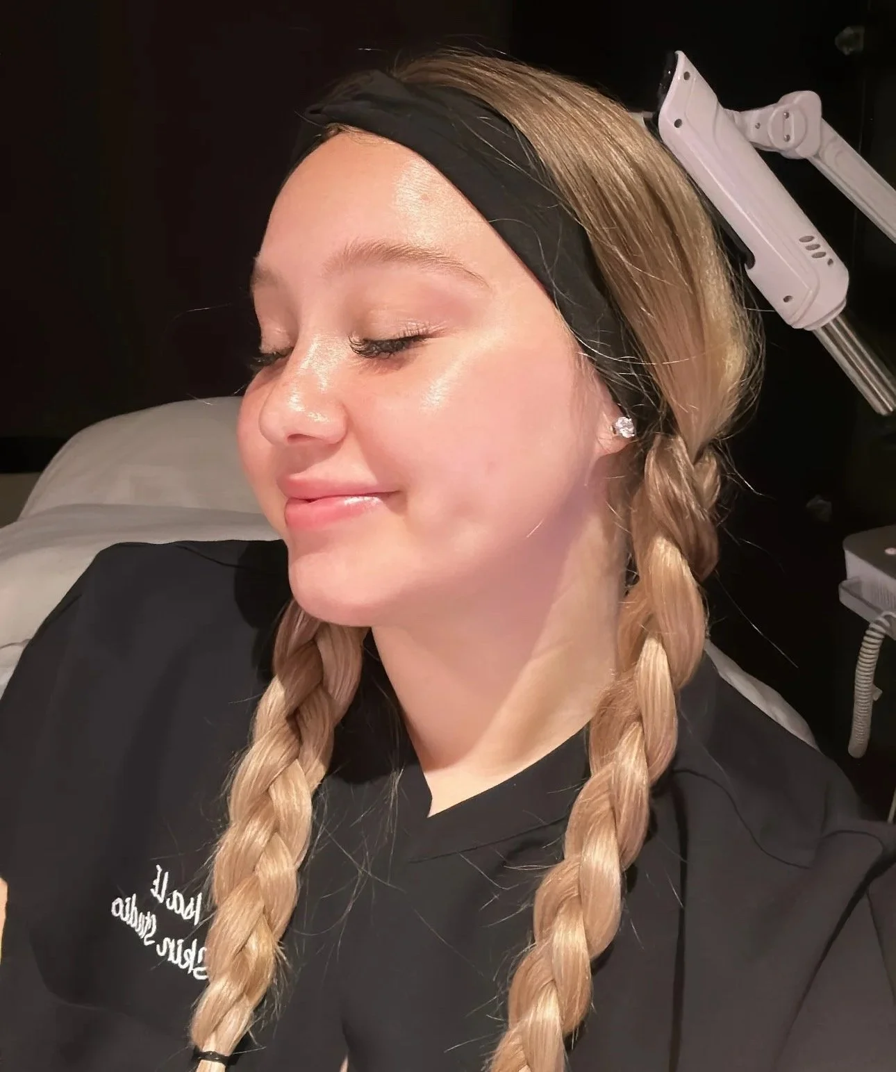 A woman with blonde hair in two braids, wearing a black headband and diamond stud earrings, lying on a bed with her eyes closed.