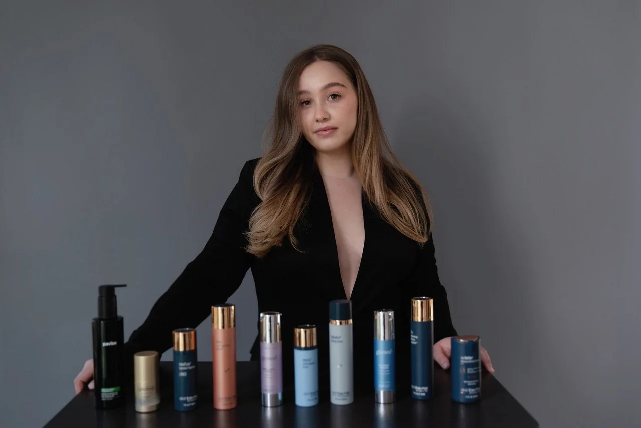 Young woman with long brown hair posing behind a makeup product display table with various skincare and cosmetic bottles and containers in front of her, against a plain gray background.